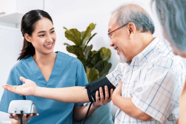 An elderly man having a blood pressure check by his personal caregiver with his wife sitting next to him in their home.