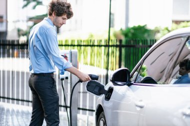 Progressive businessman insert charger plug from charging station to his electric vehicle with apartment condo building in background. Eco friendly rechargeable car powered by sustainable energy.