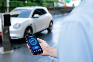 Closeup progressive businessman look at EV cars battery status application on smart phone screen at public parking charging station with power cable plug and renewable energy-powered electric vehicle