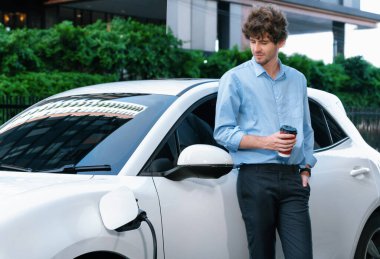 Progressive eco-friendly concept of parking EV car at public electric-powered charging station in city with blur background of businessman leaning on recharging-electric vehicle with coffee.