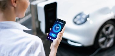 Suit-clad progressive businesswoman look at EV cars battery status from her phone while standing on a charging station with a power cable plug and a renewable energy-powered electric vehicle.