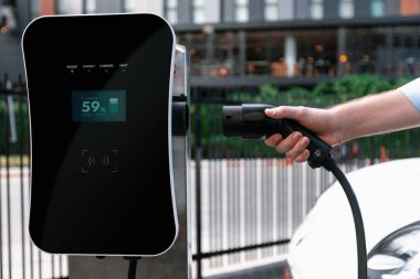 Closeup progressive man holding EV charger plug from public charging station for electric vehicle with background of residential building as concept eco-friendly sustainability energy car concept.