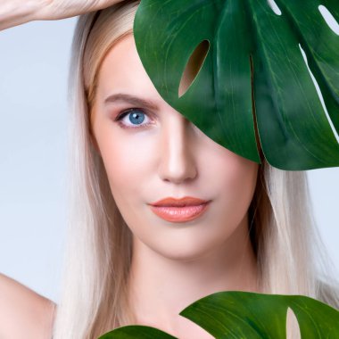 Closeup facial portrait personable woman with perfect smooth makeup holding green monstera leaves and cover her face as natural healthy skincare treatment. Tropical nature and beauty concept