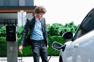 Progressive businessman wearing black suit with electric car recharging at public parking car charging station at modern city residential area. Eco friendly rechargeable car powered by clean energy.
