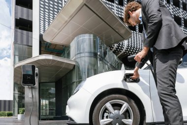 Below view closeup image of progressive black suit businessman recharge battery of his electric vehicle from public charging station. Renewable and alternative energy powered car concept.