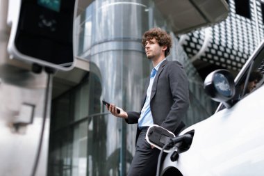 Below view of progressive businessman with electric car recharging at public charging station at modern city residential building. Eco friendly rechargeable EV car powered by alternative clean energy.