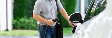 Progressive asian man install cable plug to his electric car with home charging station in the backyard. Concept use of electric vehicles in a progressive lifestyle contributes to clean environment.