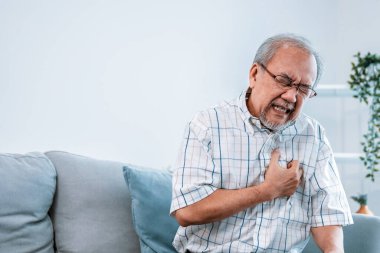 An agonizing senior man suffering from chest pain or heart attack alone in his living room. Serious health problem and feeling unwell concept for seniors.
