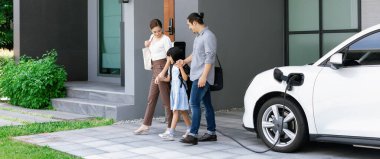 Progressive young parents and daughter with electric vehicle and home charging station. Green and clean energy from electric vehicles for healthy environment. Eco power from renewable source at home.