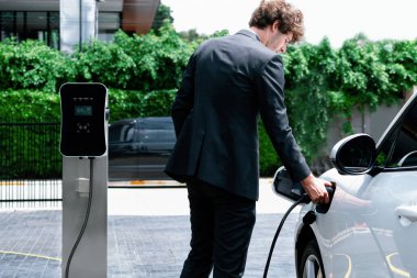 Modern şehir yerleşim bölgesindeki kamu şarj istasyonunda elektrikli araba şarj eden yenilikçi bir işadamı. Çevre dostu şarj edilebilir araba alternatif temiz enerjiyle çalışıyor..