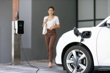 Focus image of electric vehicle recharging battery at home charging station with blurred woman walking in the background. Progressive concept of green energy technology applied in daily lifestyle.