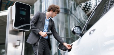 Below view closeup image of progressive black suit businessman recharge battery of his electric vehicle from public charging station. Renewable and alternative energy powered car concept.