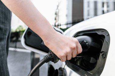 Focus EV charger plug and electric car at public charging station with blur progressive businesswoman holding charger and apartment condo building in background. Eco friendly electric vehicle concept.