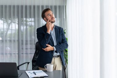 Siyah takım elbiseli yakışıklı iş adamı, modern ofis portresine güvenerek duruyor. Düşünceli bir ifadeyle iş hakkında derin düşüncelere dalmış bir sonraki hamlesini stratejik olarak düşünüyor. Varlık