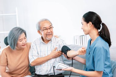 An elderly man having a blood pressure check by his personal caregiver with his wife sitting next to him in their home.