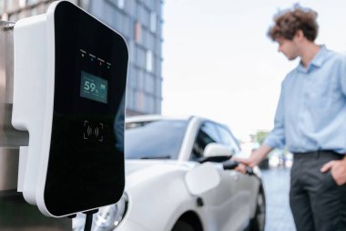 Focus public EV charging station in city residential area with blur background of businessman charging electric vehicle. Progressive lifestyle of ecological concern of city-dwelling individual.