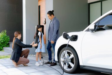 Progressive young parents and daughter with electric vehicle and home charging station. Green and clean energy from electric vehicles for healthy environment. Eco power from renewable source at home.