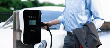 Closeup progressive suit-clad businessman with his electric vehicle recharge his car on public charging station in modern city with power cable plug and renewable energy-powered electric vehicle.