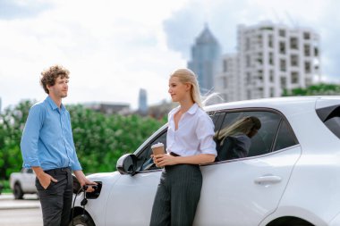 İlerici iş adamı ve kahveli iş kadını, şehir merkezinde dolaşmadan önce şarj istasyonuna bağlı elektrikli arabada duruyorlar. Çevre dostu şarj edilebilir araba temiz enerjiyle çalışıyor..