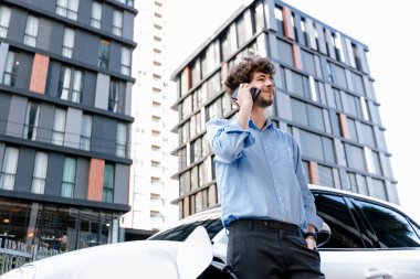 Progressive businessman talking on the phone, leaning on electric car recharging with public EV charging station, apartment condo residential building on the background as green city lifestyle.