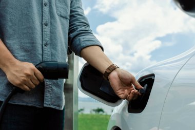 Progressive natural scenic where hand insert charging plug to electric vehicle from charging station with natural background. Future energy concept of EV car powered by sustainable electric energy.