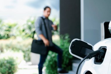 Focus electric car charging at home charging station with blurred progressive man walking in the background. Electric car using renewable clean for eco-friendly concept.