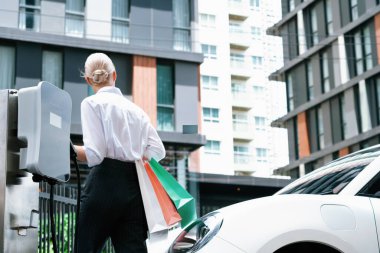 Şarj istasyonundaki elektrikli aracı alışveriş çantası taşıyan iş kadınıyla doldurmak. Temiz elektrik enerjisiyle çalışan ideal bir araba için ekolojik kaygısı olan bir adamın ilerici yaşam tarzı..