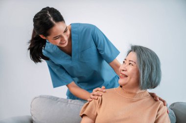 A caregiver rest her hands on the shoulders of a contented senior patient while she sitting on the sofa at home.