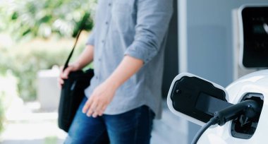 Focus electric car charging at home charging station with blurred progressive man walking in the background. Electric car using renewable clean for eco-friendly concept.