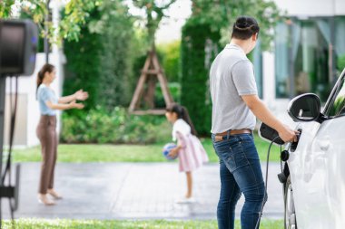 Focus image of progressive man charging electric car from home charging station with blur mother and daughter playing together in the background.