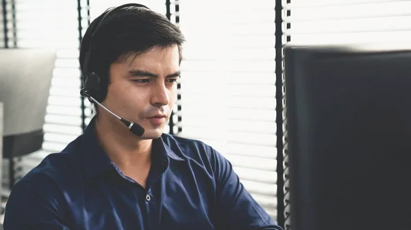 stock image Young competent asian male call center agent working at his computer while simultaneously speaking with customers. Concept of an operator, customer service agent working in the office with headset.