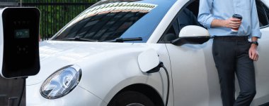 Progressive eco-friendly concept of parking EV car at public electric-powered charging station in city with blur background of businessman leaning on recharging-electric vehicle with coffee.