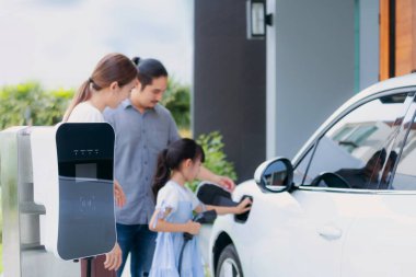 Closeup focus hand insert EV charger plug to electric vehicle at home charging station with blurred family in background. Progressive concept of green and clean energy to reduce CO2 emission by EV car