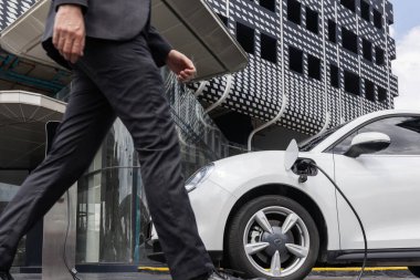 Closeup progressive businessman with electric car recharging at public charging station with background of city residential building. Eco friendly car powered by alternative clean energy.