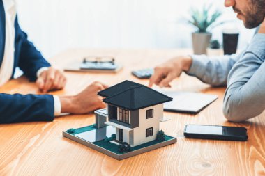 Ev modeli ahşap toplantı masasında, emlakçı ve müşterinin bulanık geçmişinde ev kredisi veya kira kontratının şartlarını tartışırken görüntülenir. Varlık