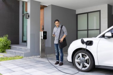 Progressive asian man and electric car with home charging station. Concept of the use of electric vehicles in a progressive lifestyle contributes to a clean and healthy environment.