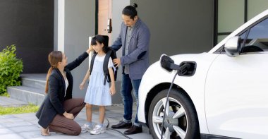 Progressive young parents and daughter with electric vehicle and home charging station. Green and clean energy from electric vehicles for healthy environment. Eco power from renewable source at home.