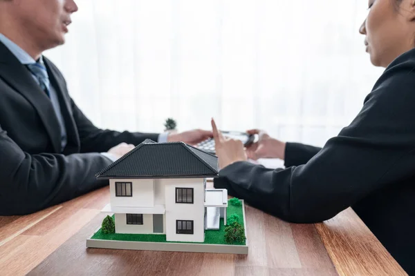 House model sample on wooden table with blurred background of real estate agent and buyer or client discussing terms and condition on house loan contract. Housing business meeting. Jubilant
