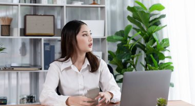 Modern ve genç hevesli iş kadını iş için akıllı telefonunu kontrol ederek ve ofisteki masasında dizüstü bilgisayarla çalışarak birden fazla iş yapıyor..