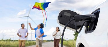 Concept of progressive happy family holding windmill toy and relax at wind farm with electric vehicle. Electric vehicle driven by clean renewable energy from wind turbine generator to charger station.