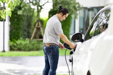 Progressive asian man install cable plug to his electric car with home charging station in the backyard. Concept use of electric vehicles in a progressive lifestyle contributes to clean environment.