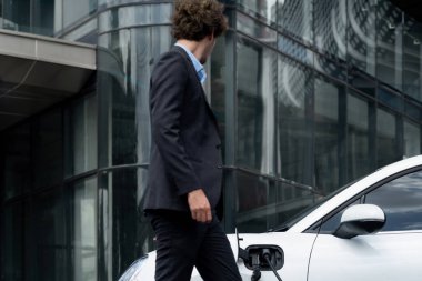 Below view of progressive businessman with electric car recharging at public charging station at modern city residential building. Eco friendly rechargeable EV car powered by alternative clean energy.