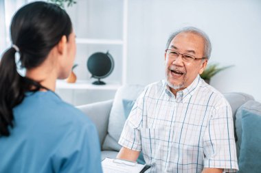Caring young female doctor examining her contented senior patient with stethoscope in living room. Medical service for elderly, elderly sickness, declining health.