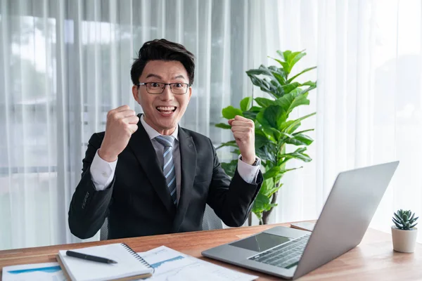 Excited Asian businessman celebrates success at office desk. Happy office worker achieves business goals with data analysis or marketing planning. Business winner celebratory expression. Jubilant