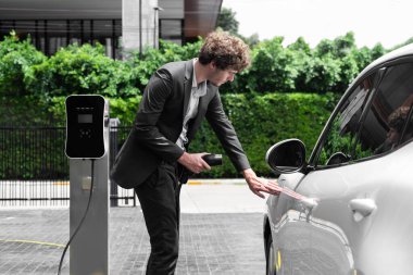 Modern şehir yerleşim bölgesindeki kamu şarj istasyonunda elektrikli araba şarj eden yenilikçi bir işadamı. Çevre dostu şarj edilebilir araba alternatif temiz enerjiyle çalışıyor..