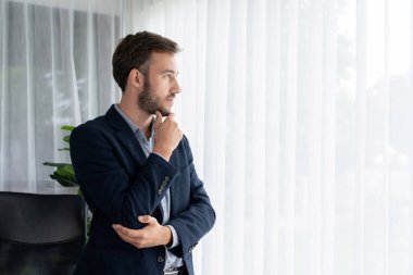 Siyah takım elbiseli yakışıklı iş adamı, modern ofis portresine güvenerek duruyor. Düşünceli bir ifadeyle iş hakkında derin düşüncelere dalmış bir sonraki hamlesini stratejik olarak düşünüyor. Varlık