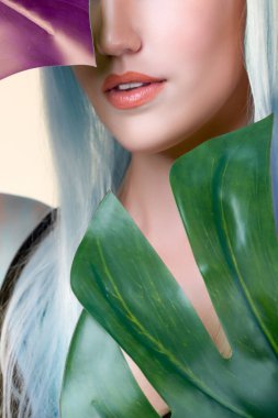 Closeup facial portrait personable woman with perfect smooth makeup holding green monstera leaves and cover her face as natural healthy skincare treatment. Tropical nature and beauty concept