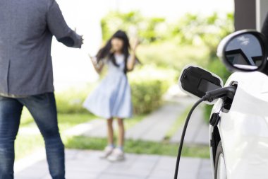 Progressive father and daughter plugs EV charger from home charging station to electric vehicle. Future eco-friendly car with EV cars powered by renewable source of clean energy.