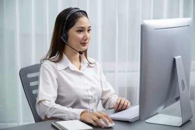 Bilgisayarında mikrofon ve kulaklıkla Asyalı çağrı merkezi çalışıyor. Kadın operatör istisnai müşteri hizmetleri sunar. Destek veren çağrı merkezi ajanı, müşteriye soruşturmada yardım ediyor. Coşkulu.