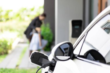 Focus EV car recharging at home charging station with blurred progressive woman and young girl in background for alternative clean energy technology concept for renewable electric vehicle.